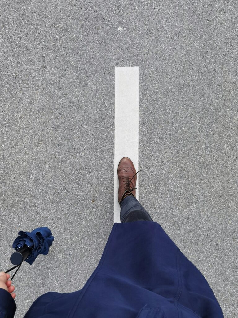 A person in a blue coat with brown boots walking on a road line, holding an umbrella.
